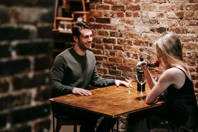 A man and woman smiling across a restaurant table as the woman holds a red rose.